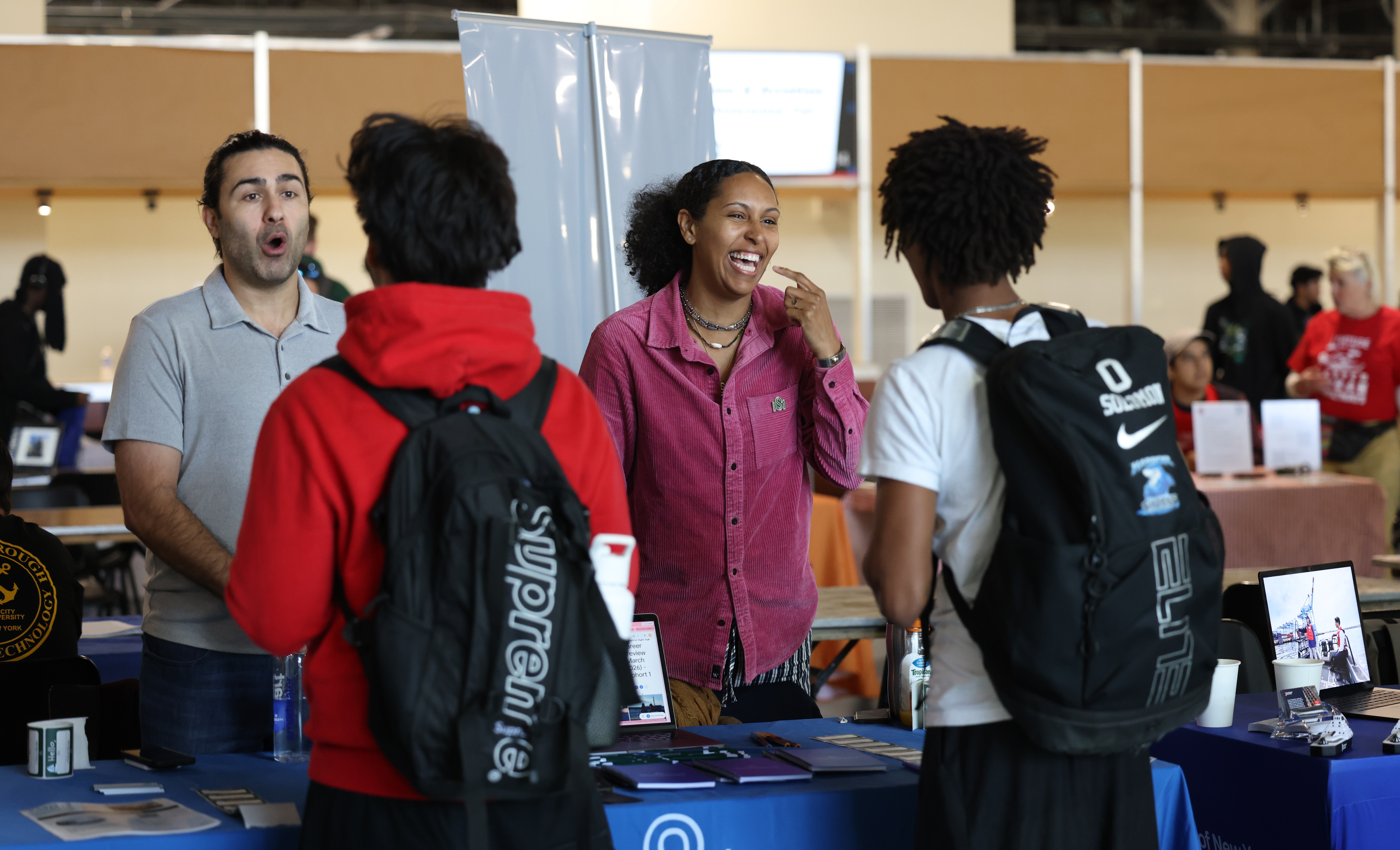 Two people stand behind a booth, smiling and talking with two students wearing backpacks at the Maritime and Aviation Career Awareness Fair, with tables and informational displays.