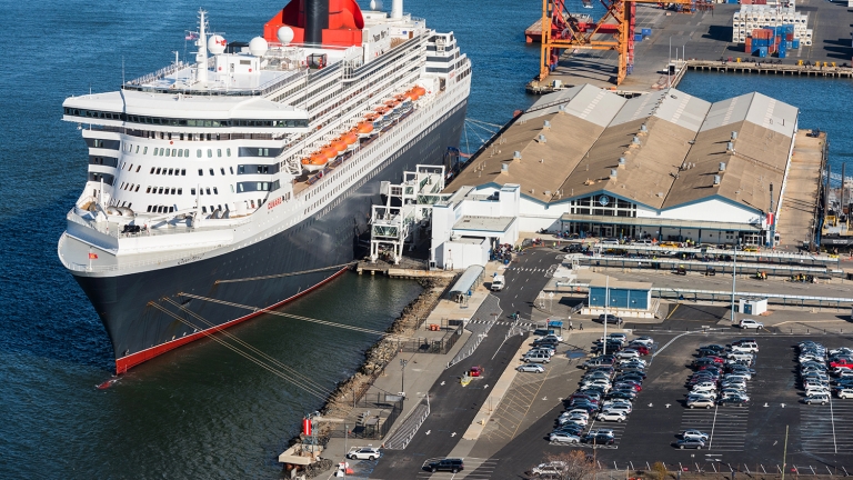 Aerial view of a large cruise ship docked at a port. The ship is white with a red funnel and multiple lifeboats along its side. Nearby are warehouses, cranes, and a parking lot filled with cars. The water is calm and blue.
