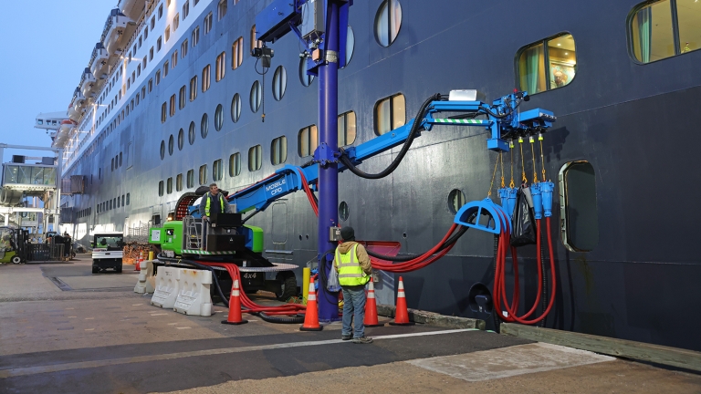 A worker in a safety vest stands near a large, blue mechanical arm connected to a cruise ship docked at the Brooklyn Cruise Terminal, with hoses attached to the ships hull. Orange cones and barriers are on the ground.