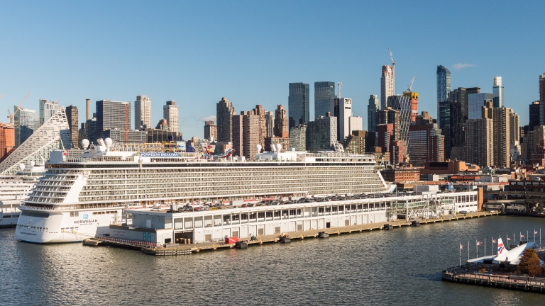 Aerial view of a large cruise ship docked at a port with a New York City skyline of high-rise buildings in the background under a clear blue sky. Photo by C. Taylor Crothers/NYCEDC.