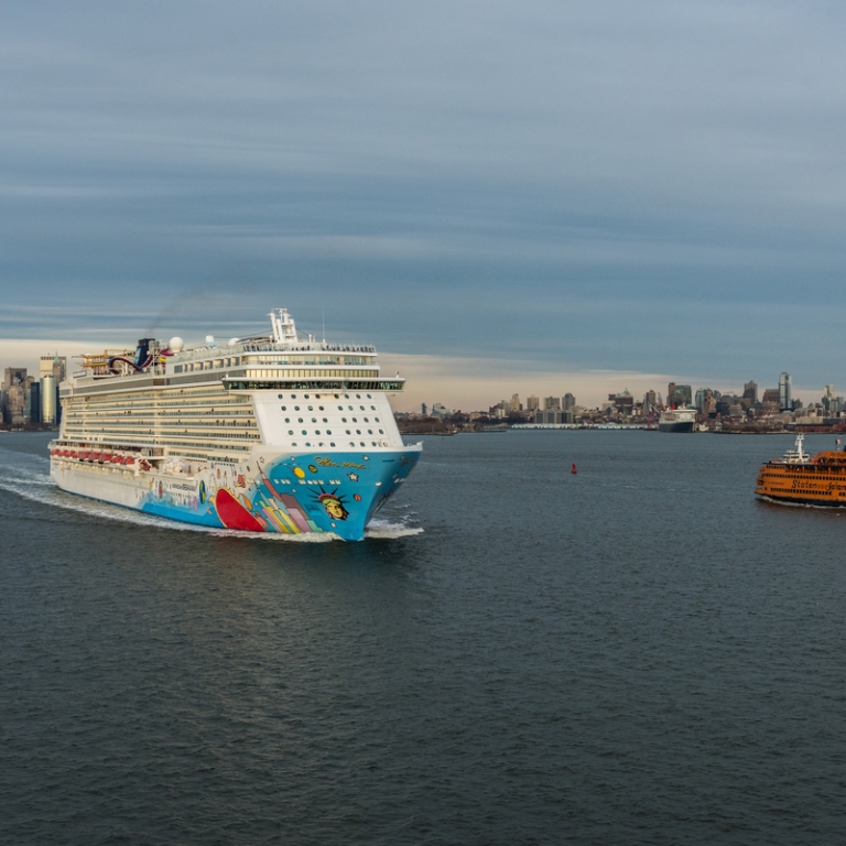 Aerial image of a large, colorful cruise ship and an orange Staten Island Ferry traveling on a river with the New York City skyline, including One World Trade Center, visible under a cloudy sky in the background. Photo courtesy of C. Taylor Crothers.