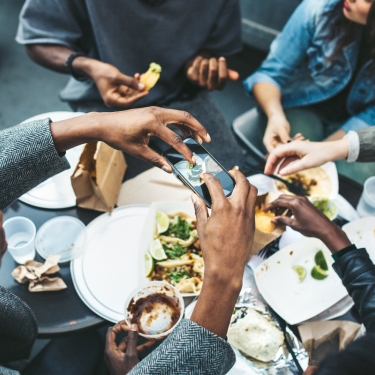 A multi-ethnic group of friends enjoy tacos and chips at a Mexican food cart in downtown New York City, New York. They laugh and talk while eating their food, having fun sharing life and culture. One of them takes a picture of their food with his smartphone to share on his social networks.