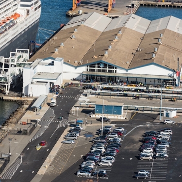 A large cruise ship is docked at Brooklyn Cruise Terminal with covered walkways connecting to the building. Numerous cars are parked in a lot nearby, and people are visible near the terminal entrance. Photo by C. Taylor Crothers.