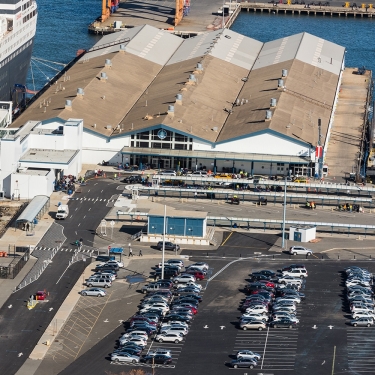 A large cruise ship is docked at Brooklyn Cruise Terminal with covered walkways connecting to the building. Numerous cars are parked in a lot nearby, and people are visible near the terminal entrance. Photo by C. Taylor Crothers.
