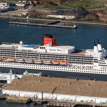 A large cruise ship, the Queen Mary 2, is docked at Brooklyn Cruise Terminal next to warehouses, with cars parked nearby and water surrounding the area. Photo by C. Taylor Crothers.