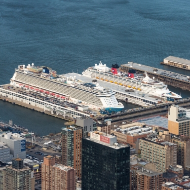 Aerial view of two large cruise ships docked at Manhattan Cruise Terminal next to tall buildings in a city, with the blue water of the harbor visible in the background. Photo by C. Taylor Crothers.