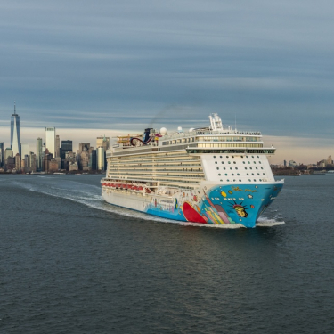 Aerial image of a large, colorful cruise ship and an orange Staten Island Ferry traveling on a river with the New York City skyline, including One World Trade Center, visible under a cloudy sky in the background. Photo courtesy of C. Taylor Crothers.