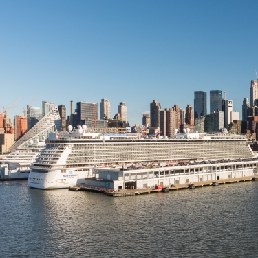 A large white cruise ship docked at Manhattan Cruise Terminal with the New York City skyline and skyscrapers in the background on a clear, sunny day. Photo by C. Taylor Crothers.