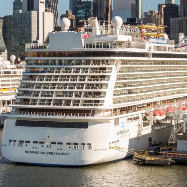 A large white cruise ship, Norwegian Breakaway, docked at a busy Manhattan Cruise Terminal with NYC buildings in the background. Photo by C. Taylor Crothers.