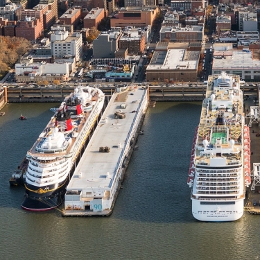 Aerial view of two large cruise ships docked at Manhattan Cruise Terminal, with numerous parked cars, nearby buildings, and a green park visible in the background. Photo by C. Taylor Crothers.