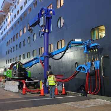 A worker in a safety vest stands near a large, blue mechanical arm connected to a cruise ship docked at the Brooklyn Cruise Terminal, with hoses attached to the ships hull. Orange cones and barriers are on the ground.