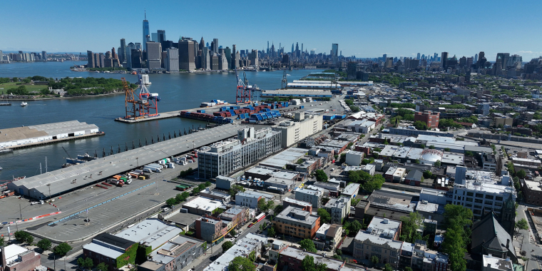 Aerial view of Brooklyn Marine Terminal with shipping containers and cranes beside an urban area; the Manhattan skyline and waterfront are visible across the river under a clear blue sky.