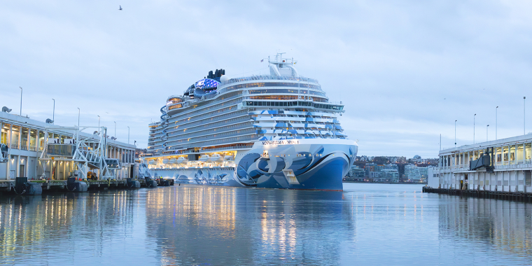 A large cruise ship, the Norwegian Prima, between two piers at the Manhattan Cruise Terminal on a calm, cloudy day. The ship features blue and white designs on its bow and is illuminated with lights. Buildings are visible in the background.