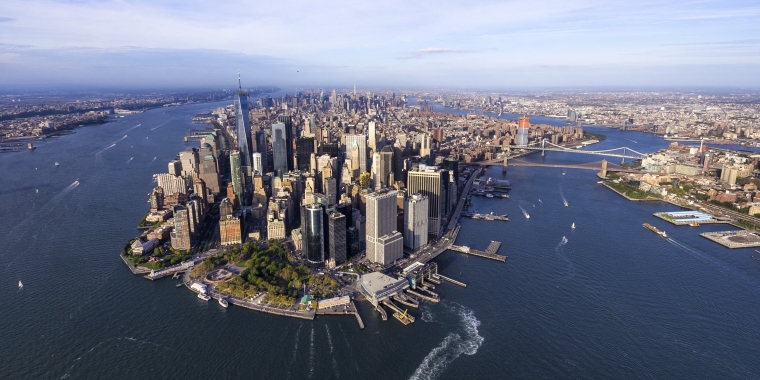 Aerial view of Lower Manhattan, New York City, showing skyscrapers, waterfront parks, and surrounding rivers with boats. The Brooklyn and Manhattan bridges are visible in the background. Photo by Eloi Omella/Getty Images.
