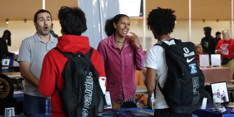 Two people stand behind a booth, smiling and talking with two students wearing backpacks at the Maritime and Aviation Career Awareness Fair, with tables and informational displays.
